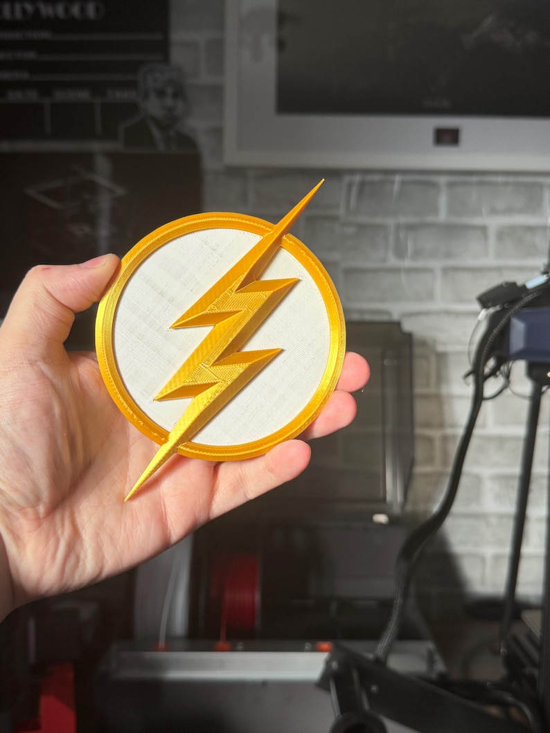 May include: A gold-colored lightning bolt emblem, set against a white circle, is framed by a gold border. The emblem is held in a hand, showcasing the 3D-printed design. The background is blurred, suggesting a workshop setting.