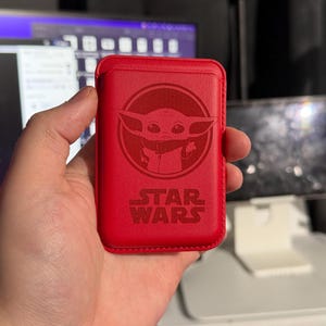 May include: A red leather phone case featuring a circular embossed image of Grogu, also known as Baby Yoda, from Star Wars. The Star Wars logo is below the image. The case is rectangular and appears to be a wallet-style design.