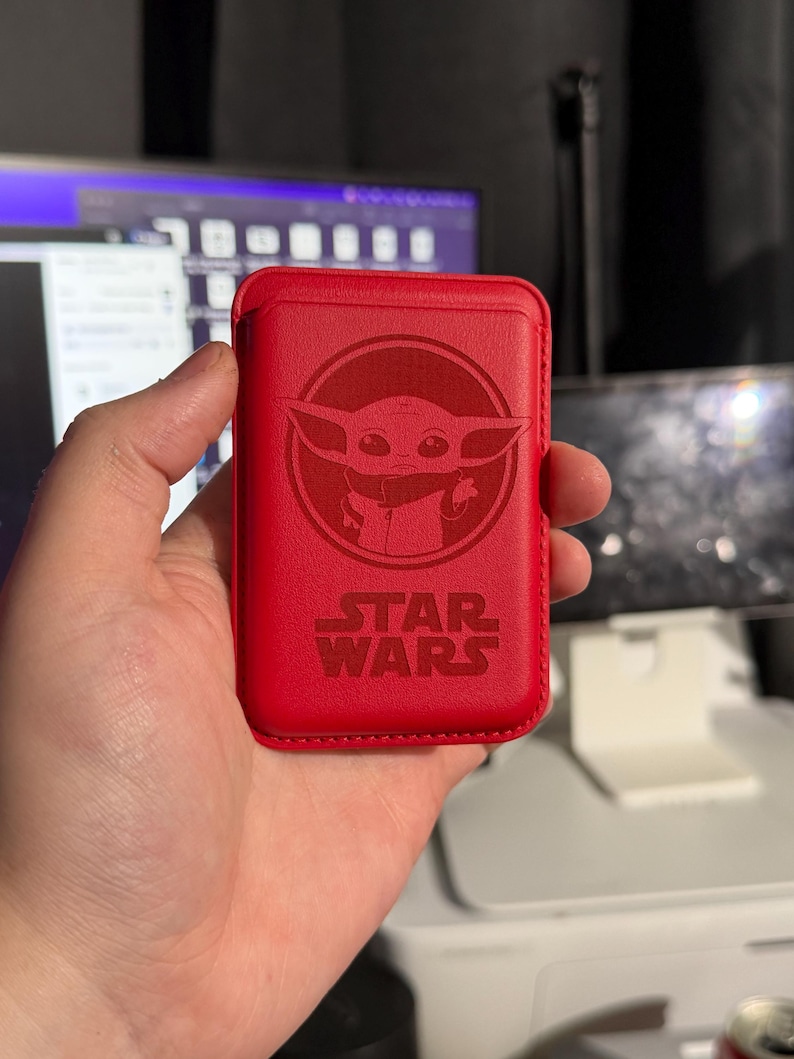 May include: A red leather phone case featuring a circular embossed image of Grogu, also known as Baby Yoda, from Star Wars. The Star Wars logo is below the image. The case is rectangular and appears to be a wallet-style design.