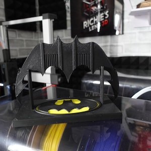 May include: A black 3D printed display stand featuring a bat-shaped archway and a base with a yellow and black bat logo emblem. The structure is composed of a rectangular platform supporting a stylized bat wing silhouette with vertical detailing.