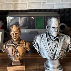 May include: Two 3D-printed busts of comic book characters. The bust on the left is bronze-colored and features a character with horns and the letter "D". The bust on the right is silver and depicts a man in a suit.