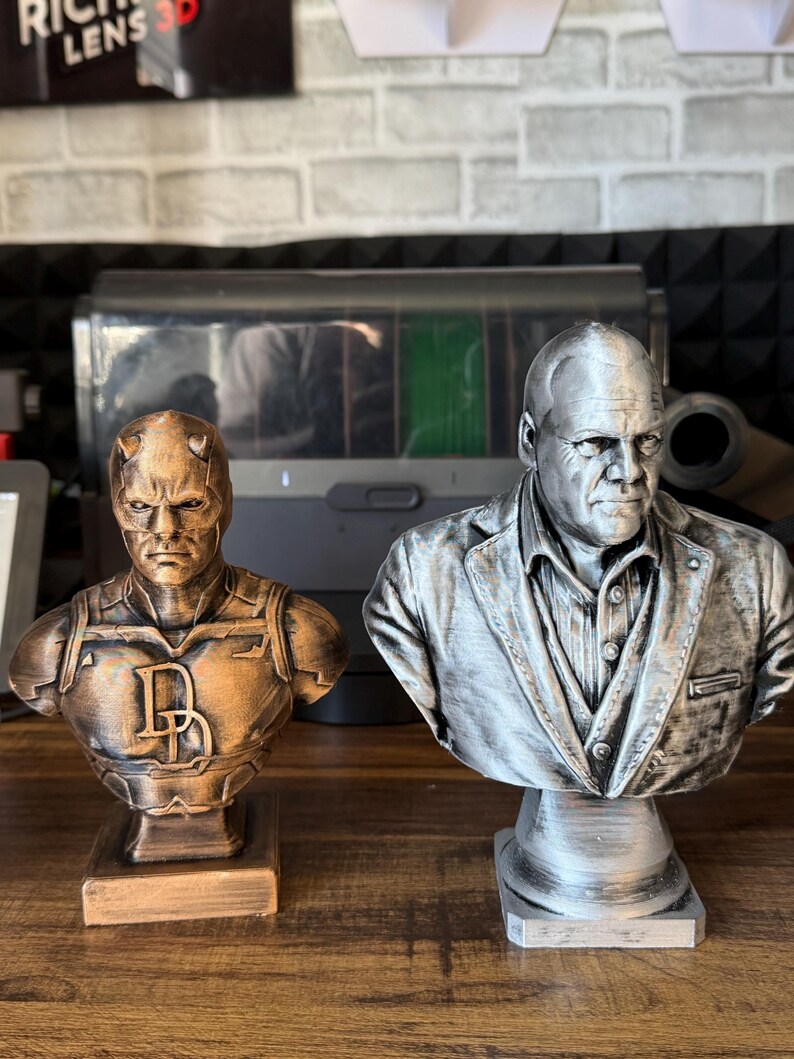 May include: Two 3D-printed busts of comic book characters. The bust on the left is bronze-colored and features a character with horns and the letter "D". The bust on the right is silver and depicts a man in a suit.