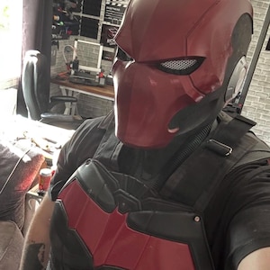 May include: A Red Hood helmet and chest armour set. The helmet is a deep red with a matte finish, featuring a mesh eye covering. The chest armour is black with a red bat symbol on the chest. The overall design is reminiscent of a comic book superhero.