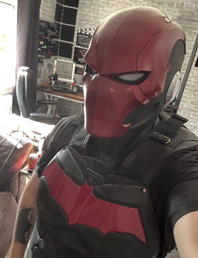 May include: A Red Hood helmet and chest armour set. The helmet is a deep red with a matte finish, featuring a mesh eye covering. The chest armour is black with a red bat symbol on the chest. The overall design is reminiscent of a comic book superhero.