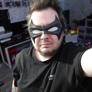 May include: A black superhero-style eye mask featuring a pointed, angular design with white mesh inserts over the eye openings. The mask covers the upper face and is constructed from a solid, matte-textured material with distinct bat-like features.