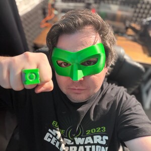 Puede incluir: Se muestra una m&aacute;scara y un anillo de superh&eacute;roe verdes. La m&aacute;scara cubre los ojos y la nariz, y el anillo est&aacute; en un dedo. El anillo y la m&aacute;scara son de color verde brillante. La persona lleva una camiseta negra con el texto "ELITE 2023 STAR WARS CELEBRATION".