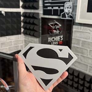 May include: A diamond-shaped emblem featuring a stylized letter S in light gray layered over a solid black background. The design mimics a classic superhero insignia with sharp, geometric edges and a clean, high-contrast two-tone color scheme.