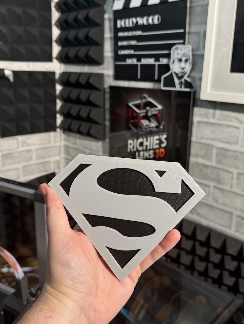 May include: A diamond-shaped emblem featuring a stylized letter S in light gray layered over a solid black background. The design mimics a classic superhero insignia with sharp, geometric edges and a clean, high-contrast two-tone color scheme.