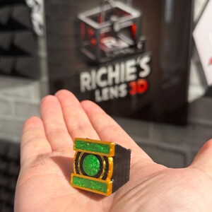 Puede incluir: Un peque&ntilde;o objeto rectangular impreso en 3D con un dise&ntilde;o verde y dorado, sostenido en una mano. El objeto tiene un centro verde circular y adornos dorados. El fondo presenta un letrero con el texto "RICHIE'S LENS 3D".