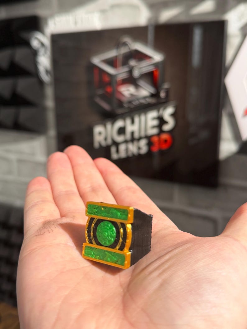 Puede incluir: Un peque&ntilde;o objeto rectangular impreso en 3D con un dise&ntilde;o verde y dorado, sostenido en una mano. El objeto tiene un centro verde circular y adornos dorados. El fondo presenta un letrero con el texto "RICHIE'S LENS 3D".
