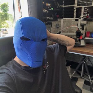 Op de afbeelding: Een blauw stoffen masker met oogopeningen, dat doet denken aan een superheld of stripfiguur. Het masker is het centrale element, met een persoon die een zwart t-shirt op de achtergrond draagt. Het masker heeft een gestructureerd oppervlak en is ontworpen om het hele hoofd te bedekken.