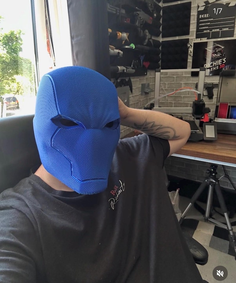 Op de afbeelding: Een blauw stoffen masker met oogopeningen, dat doet denken aan een superheld of stripfiguur. Het masker is het centrale element, met een persoon die een zwart t-shirt op de achtergrond draagt. Het masker heeft een gestructureerd oppervlak en is ontworpen om het hele hoofd te bedekken.