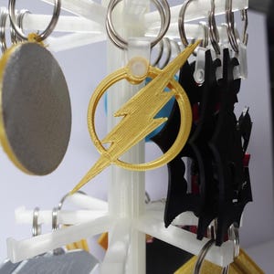 K&ouml;nnte beinhalten: Ein wei&szlig;er Displayst&auml;nder mit verschiedenen Schl&uuml;sselanh&auml;ngern. Zu den Schl&uuml;sselanh&auml;ngern geh&ouml;ren ein goldener Blitz, eine schwarze Silhouette und ein graues Oval. Jeder Schl&uuml;sselanh&auml;nger hat einen silbernen Ring.