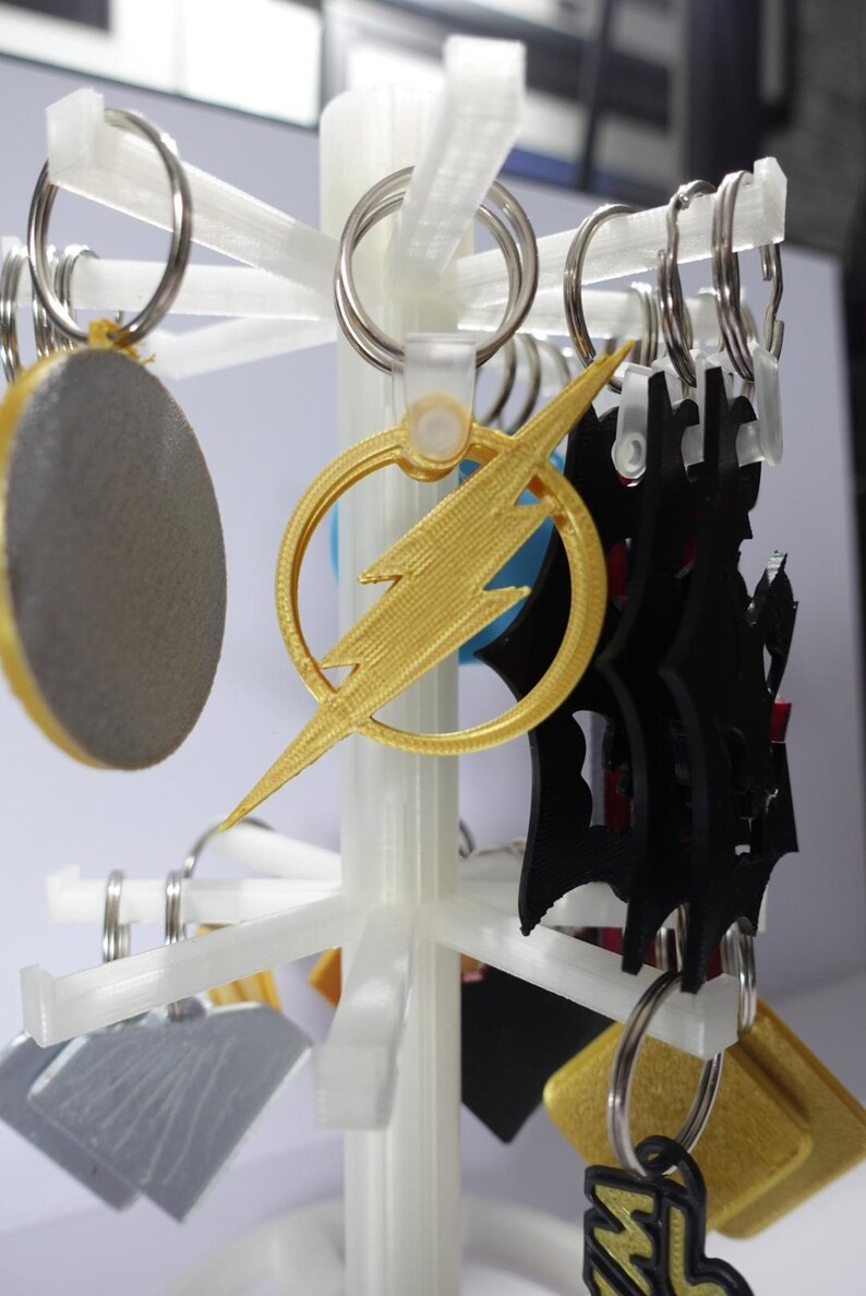 K&ouml;nnte beinhalten: Ein wei&szlig;er Displayst&auml;nder mit verschiedenen Schl&uuml;sselanh&auml;ngern. Zu den Schl&uuml;sselanh&auml;ngern geh&ouml;ren ein goldener Blitz, eine schwarze Silhouette und ein graues Oval. Jeder Schl&uuml;sselanh&auml;nger hat einen silbernen Ring.
