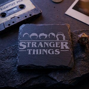 May include: A square, dark gray slate coaster with the words "STRANGER THINGS" and character silhouettes etched into the surface. A vintage cassette tape labeled "EIGHTIES MIX" and a die are in the background.