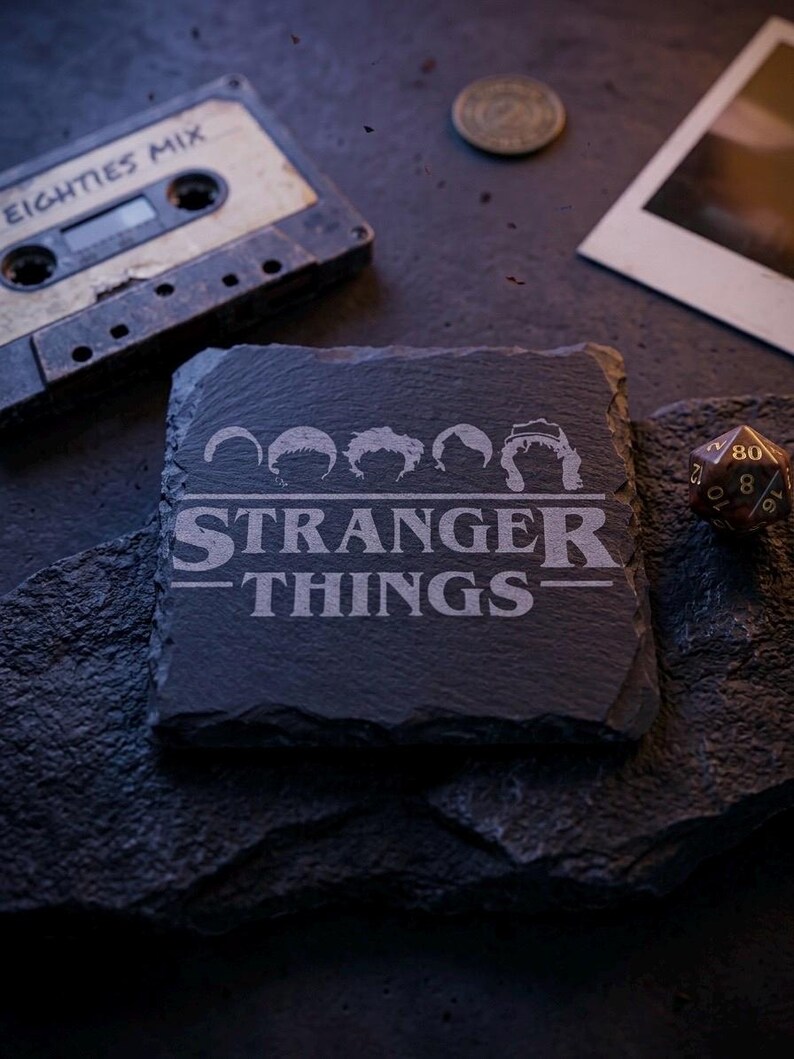 May include: A square, dark gray slate coaster with the words "STRANGER THINGS" and character silhouettes etched into the surface. A vintage cassette tape labeled "EIGHTIES MIX" and a die are in the background.