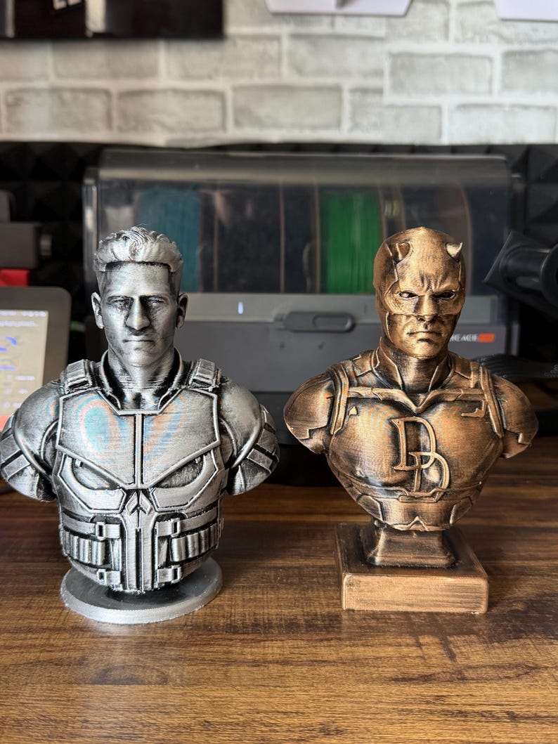 May include: Two superhero bust statues. The statue on the left is silver and depicts a character with a stern expression and a detailed chest plate. The statue on the right is bronze and features a character with a mask and the letter "D".