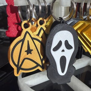 K&ouml;nnte beinhalten: Schl&uuml;sselanh&auml;nger mit Popkultur-Designs. Einer ist ein gold-schwarzes Star Trek-Emblem. Ein anderer ist eine schwarz-wei&szlig;e Ghostface-Maske. Jeder Schl&uuml;sselanh&auml;nger hat einen silbernen Metallring.