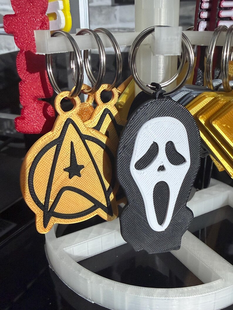 K&ouml;nnte beinhalten: Schl&uuml;sselanh&auml;nger mit Popkultur-Designs. Einer ist ein gold-schwarzes Star Trek-Emblem. Ein anderer ist eine schwarz-wei&szlig;e Ghostface-Maske. Jeder Schl&uuml;sselanh&auml;nger hat einen silbernen Metallring.