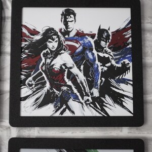 May include: A framed art print featuring a stylized illustration of Wonder Woman, Superman, and Batman. The artwork uses a high-contrast, ink-wash aesthetic with bold black lines and accents of red and blue against a white background.