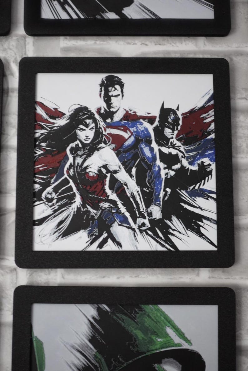 May include: A framed art print featuring a stylized illustration of Wonder Woman, Superman, and Batman. The artwork uses a high-contrast, ink-wash aesthetic with bold black lines and accents of red and blue against a white background.