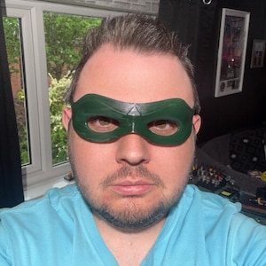 May include: A dark green domino mask with a smooth, glossy finish and a central triangular detail above the nose bridge. The mask features two wide, rounded eye openings and contoured edges that follow the shape of the upper face area.