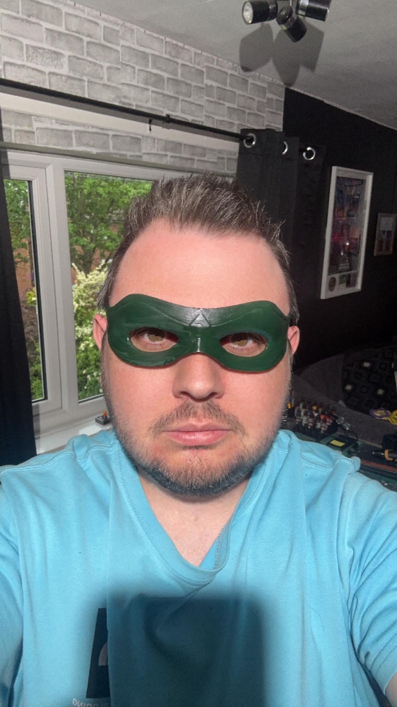 May include: A dark green domino mask with a smooth, glossy finish and a central triangular detail above the nose bridge. The mask features two wide, rounded eye openings and contoured edges that follow the shape of the upper face area.