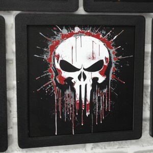 May include: A square black frame containing a graphic print of a white skull with black eye sockets and nose cavity. The design features red and grey paint splatter effects that drip downwards over a solid black background within the frame.