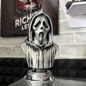 May include: A silver-colored bust of the Ghostface mask from the Scream horror film franchise. The bust features a detailed rendering of the mask's iconic expression and hooded cloak, mounted on a decorative base. The bust is displayed on a reflective surface.