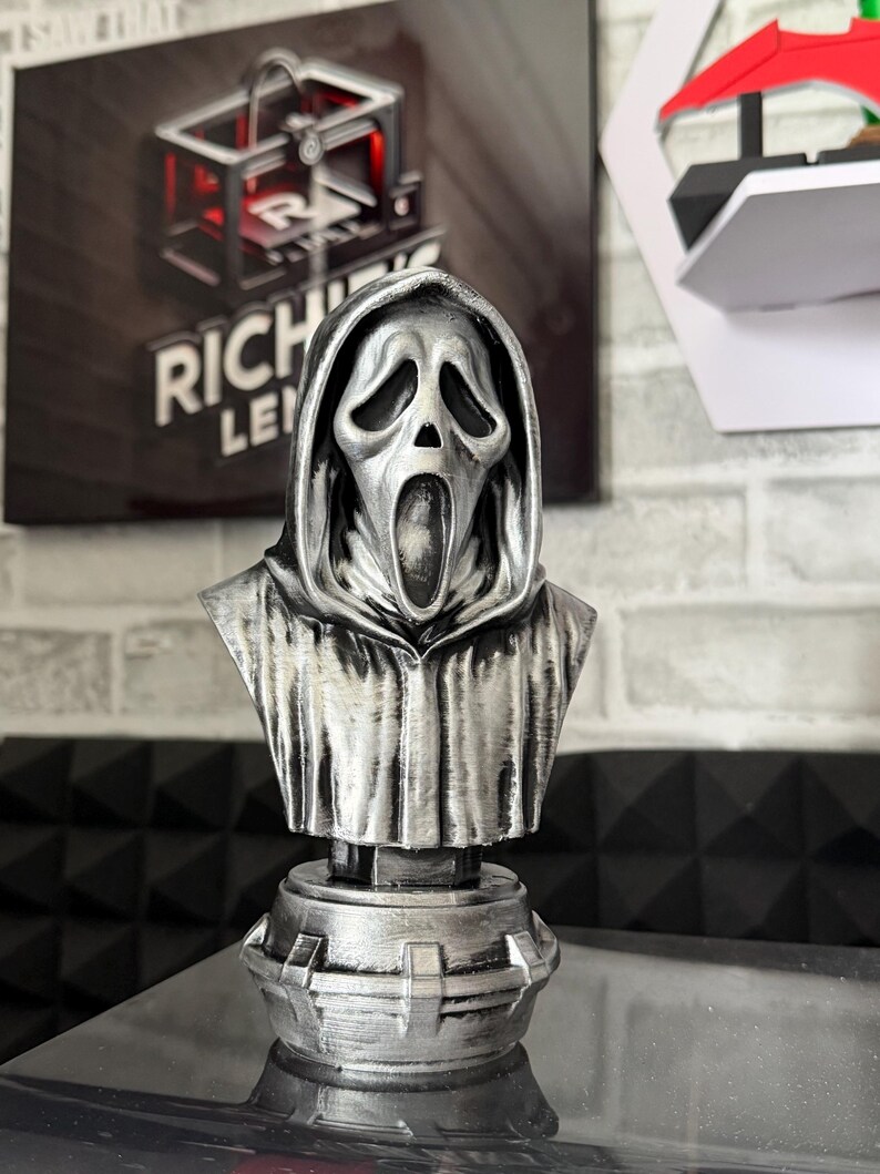 May include: A silver-colored bust of the Ghostface mask from the Scream horror film franchise. The bust features a detailed rendering of the mask's iconic expression and hooded cloak, mounted on a decorative base. The bust is displayed on a reflective surface.