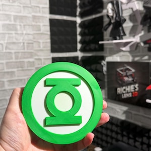 May include: A circular Green Lantern emblem featuring a bright green outer ring and a matching green central symbol set against a white background. The object has a flat, layered design with visible 3D printed texture lines across the surface.