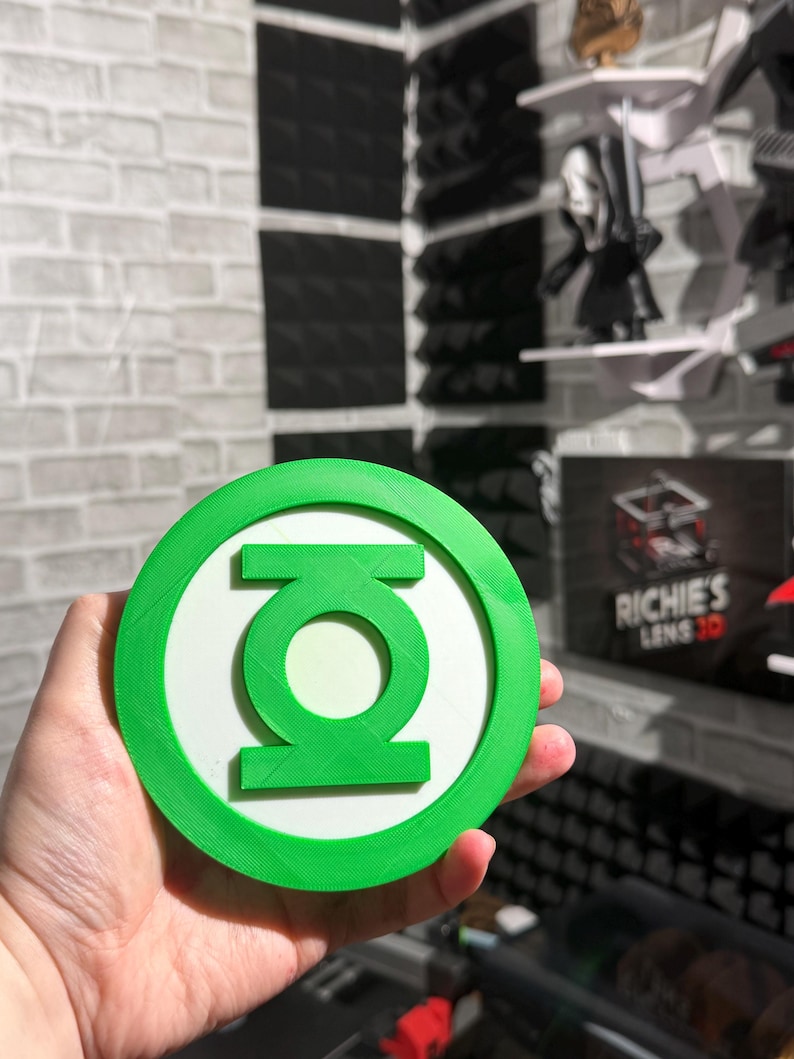 May include: A circular Green Lantern emblem featuring a bright green outer ring and a matching green central symbol set against a white background. The object has a flat, layered design with visible 3D printed texture lines across the surface.