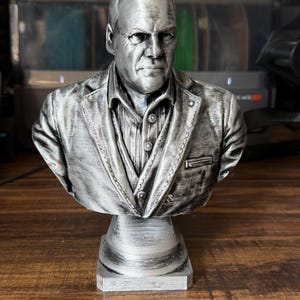 May include: A silver-colored bust of a man wearing a suit jacket and collared shirt. The bust is mounted on a square pedestal. The sculpture has detailed facial features and clothing textures.