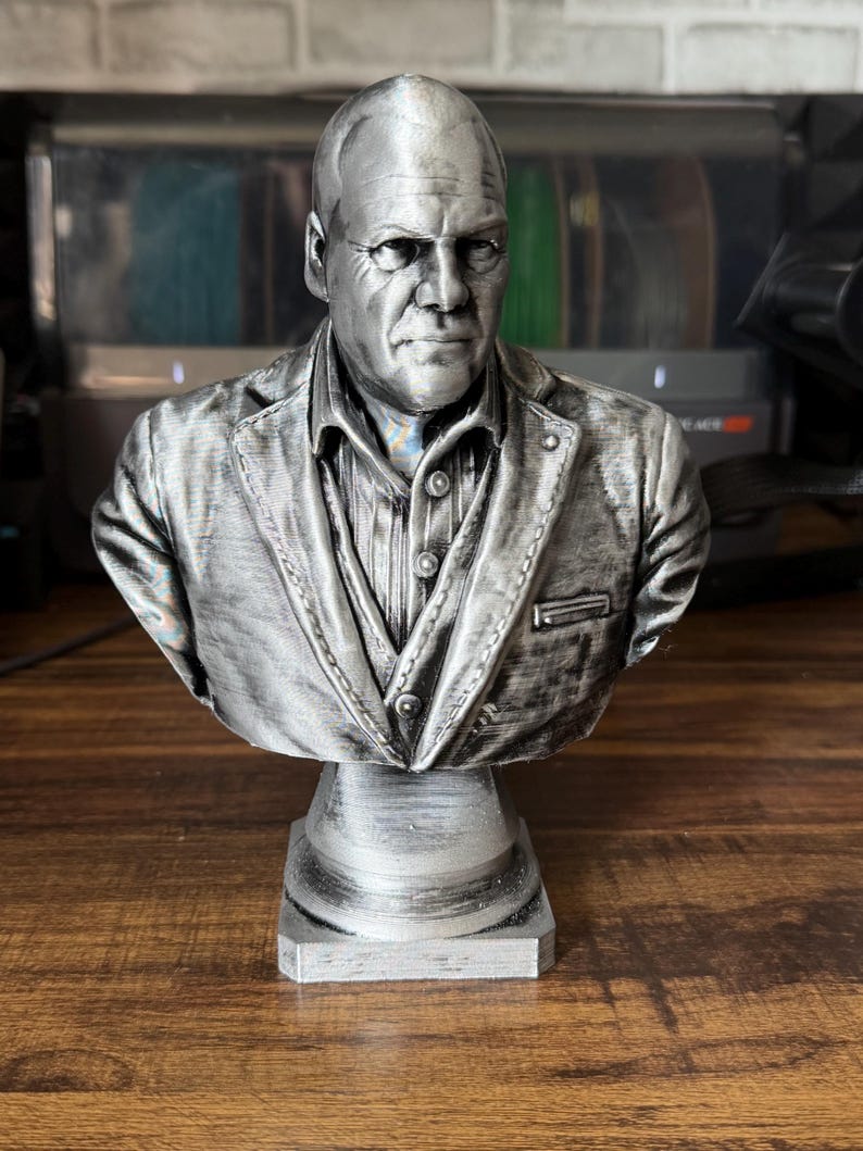 May include: A silver-colored bust of a man wearing a suit jacket and collared shirt. The bust is mounted on a square pedestal. The sculpture has detailed facial features and clothing textures.