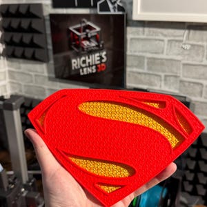 May include: A 3D printed Superman shield emblem featuring a bright red outer frame with a textured surface and a recessed yellow inner section with a detailed, repeating pattern. The iconic diamond-shaped logo is constructed with layered plastic.