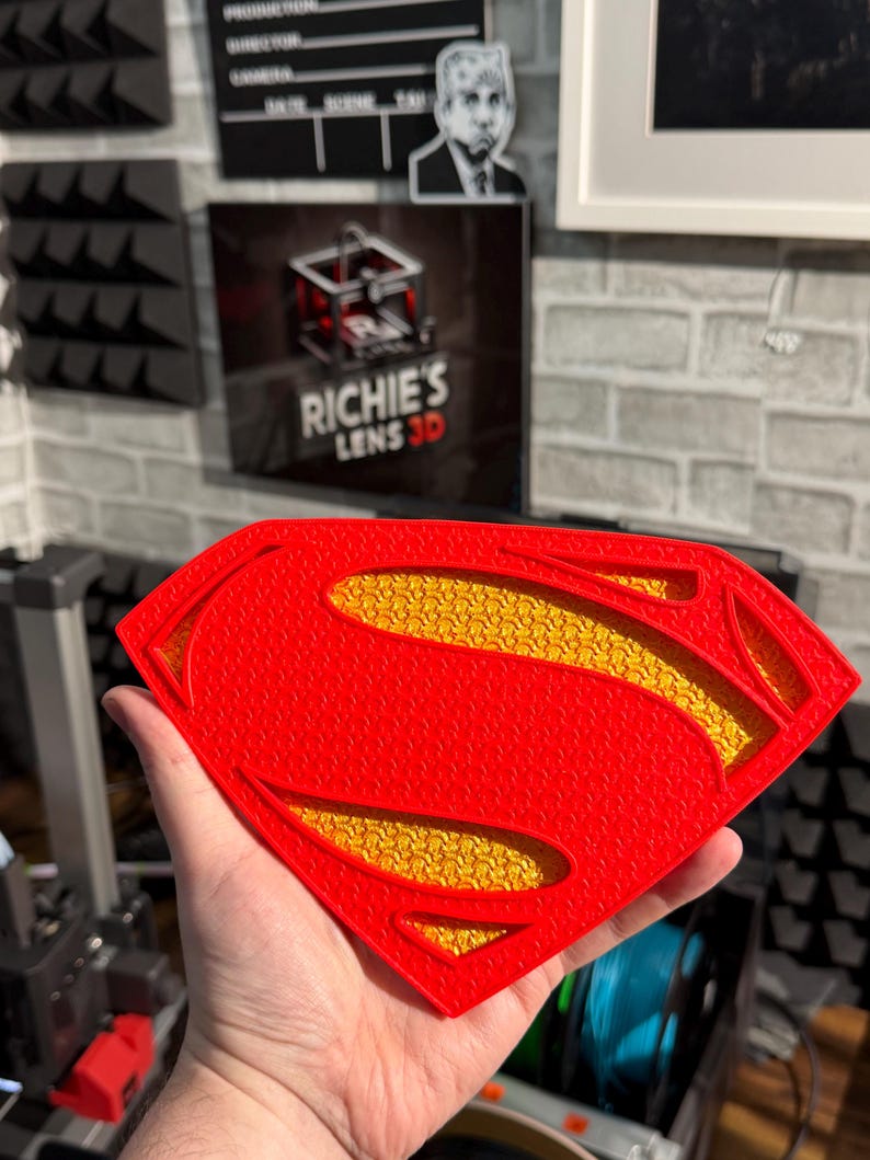May include: A 3D printed Superman shield emblem featuring a bright red outer frame with a textured surface and a recessed yellow inner section with a detailed, repeating pattern. The iconic diamond-shaped logo is constructed with layered plastic.