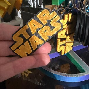 K&ouml;nnte beinhalten: Ein goldener und schwarzer Star Wars Schl&uuml;sselanh&auml;nger. Der Schl&uuml;sselanh&auml;nger zeigt das Star Wars-Logo in goldfarbenen Buchstaben auf schwarzem Hintergrund. Der Schl&uuml;sselanh&auml;nger ist an einem silbernen Schl&uuml;sselring befestigt. Weitere Schl&uuml;sselanh&auml;nger sind im Hintergrund zu sehen.