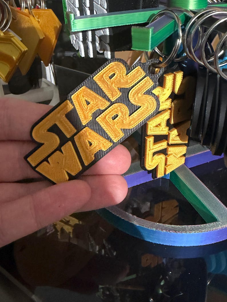 K&ouml;nnte beinhalten: Ein goldener und schwarzer Star Wars Schl&uuml;sselanh&auml;nger. Der Schl&uuml;sselanh&auml;nger zeigt das Star Wars-Logo in goldfarbenen Buchstaben auf schwarzem Hintergrund. Der Schl&uuml;sselanh&auml;nger ist an einem silbernen Schl&uuml;sselring befestigt. Weitere Schl&uuml;sselanh&auml;nger sind im Hintergrund zu sehen.
