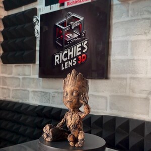 May include: A brown 3D-printed Groot figurine, seated on a black rotating base. The figurine is detailed with textured bark-like features. The background includes signs that say "Hollywood" and "Like Subscribe RichiesLens".