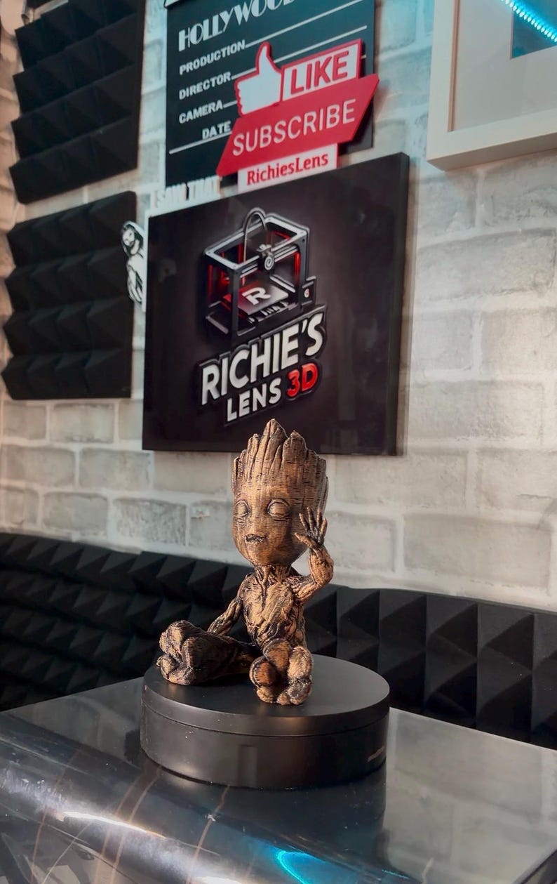 May include: A brown 3D-printed Groot figurine, seated on a black rotating base. The figurine is detailed with textured bark-like features. The background includes signs that say "Hollywood" and "Like Subscribe RichiesLens".