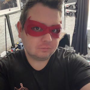 May include: A red, molded plastic eye mask with pointed outer edges that covers the upper face. The mask features a smooth, matte finish and is designed to fit around the eyes, extending toward the temples with a distinct superhero-inspired shape.