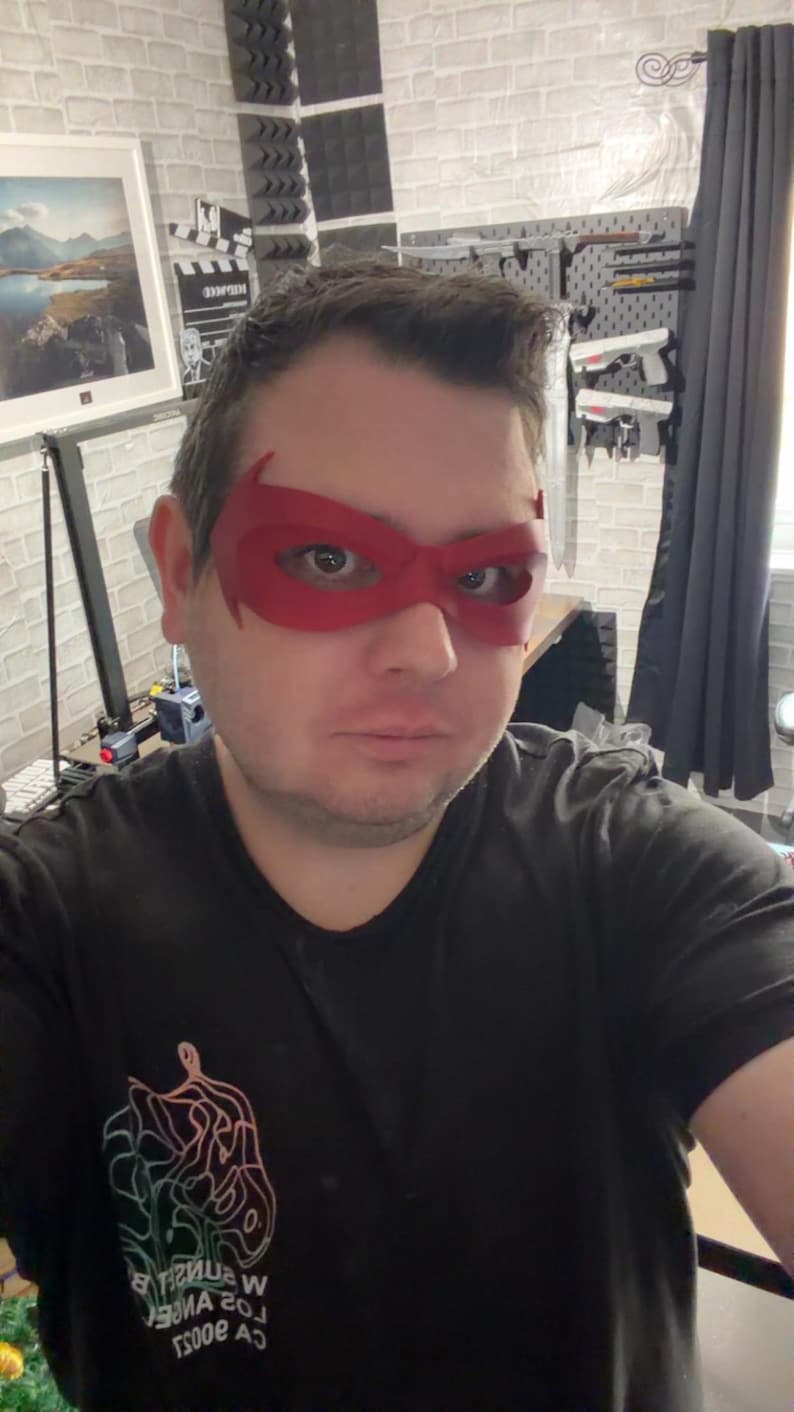 May include: A red, molded plastic eye mask with pointed outer edges that covers the upper face. The mask features a smooth, matte finish and is designed to fit around the eyes, extending toward the temples with a distinct superhero-inspired shape.