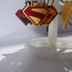 K&ouml;nnte beinhalten: Schl&uuml;sselanh&auml;nger mit dem Superman-Logo in Rot und Gelb, pr&auml;sentiert auf einem wei&szlig;en St&auml;nder. Das Logo hat die Form einer Raute mit rotem Rand und einem gelben 'S' in der Mitte. Weitere Schl&uuml;sselanh&auml;nger mit verschiedenen Designs sind ebenfalls sichtbar.
