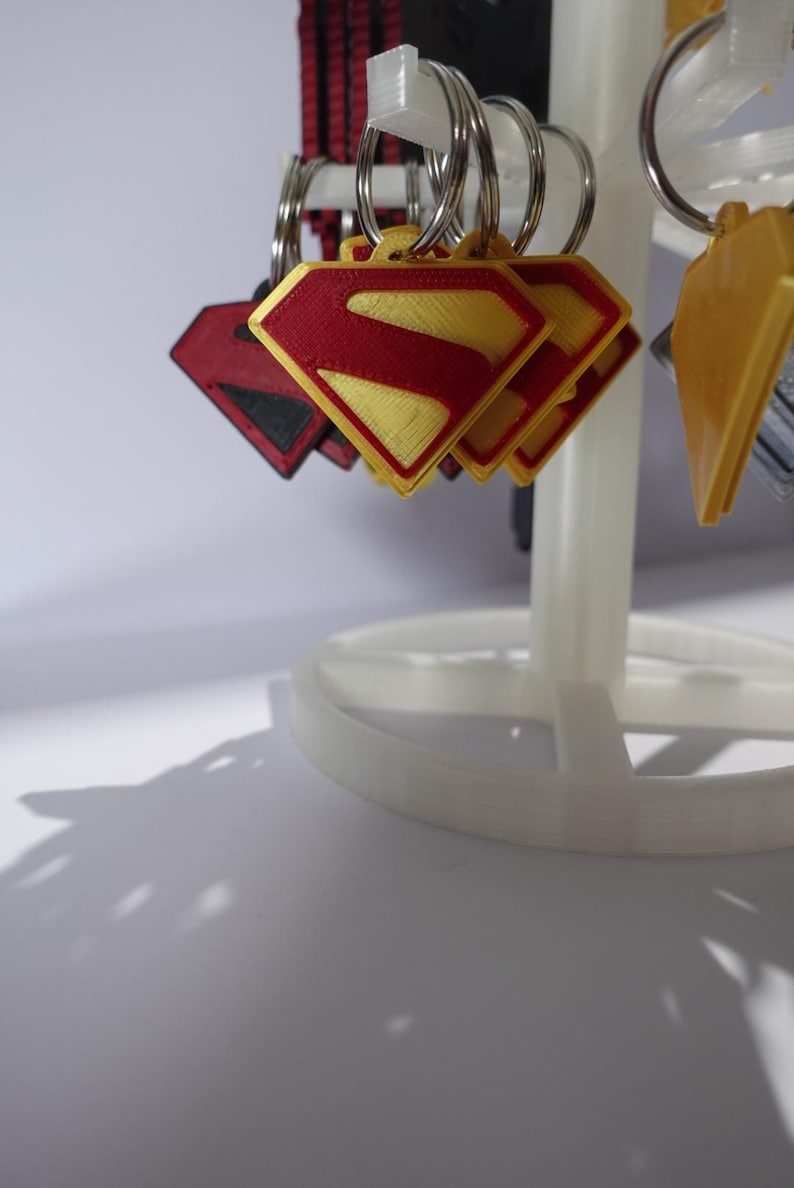 K&ouml;nnte beinhalten: Schl&uuml;sselanh&auml;nger mit dem Superman-Logo in Rot und Gelb, pr&auml;sentiert auf einem wei&szlig;en St&auml;nder. Das Logo hat die Form einer Raute mit rotem Rand und einem gelben 'S' in der Mitte. Weitere Schl&uuml;sselanh&auml;nger mit verschiedenen Designs sind ebenfalls sichtbar.