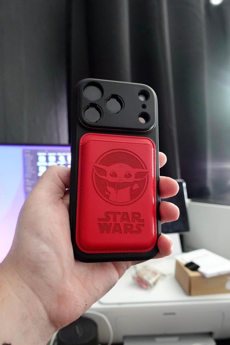 May include: Black phone case with a red magnetic card holder featuring a Baby Yoda design and the Star Wars logo. The case has a black camera module with three lenses.