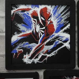 May include: A square framed wall art piece featuring a stylized illustration of a superhero in a red and blue suit with a white spider emblem on the chest. The character is depicted in a dynamic pose against a black background with white paint splatter effects.