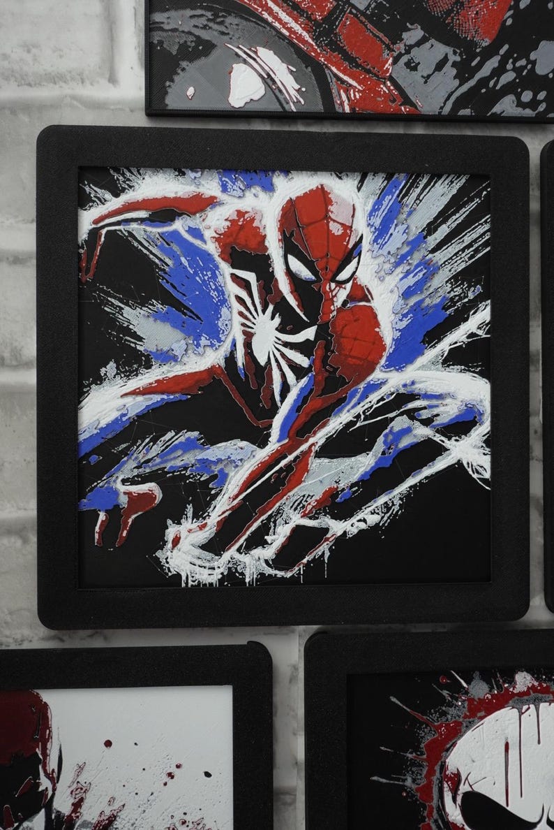 May include: A square framed wall art piece featuring a stylized illustration of a superhero in a red and blue suit with a white spider emblem on the chest. The character is depicted in a dynamic pose against a black background with white paint splatter effects.