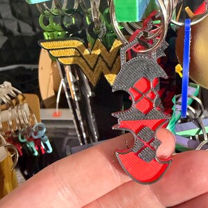 K&ouml;nnte beinhalten: Eine Sammlung von Schl&uuml;sselanh&auml;ngern, darunter ein rot-schwarzer Charm in Axtform, ein gold-gelbes Wonder Woman-Logo und andere farbenfrohe Designs. Die Schl&uuml;sselanh&auml;nger sind an silbernen Metallringen befestigt.