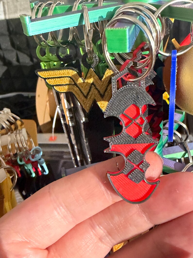 K&ouml;nnte beinhalten: Eine Sammlung von Schl&uuml;sselanh&auml;ngern, darunter ein rot-schwarzer Charm in Axtform, ein gold-gelbes Wonder Woman-Logo und andere farbenfrohe Designs. Die Schl&uuml;sselanh&auml;nger sind an silbernen Metallringen befestigt.