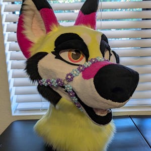 May include: A yellow, black, and pink furry wolf head with a beaded nose chain. The ears are black and pink, and the eyes are orange and black. The muzzle is black, and the fur is yellow. The head is designed for cosplay or costume use.